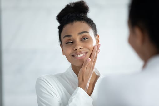 Head shot smiling African American woman touching cheek, enjoying perfect smooth face skin, looking in mirror, beautiful girl wearing white bathrobe doing facial massage in bathroom, skincare concept