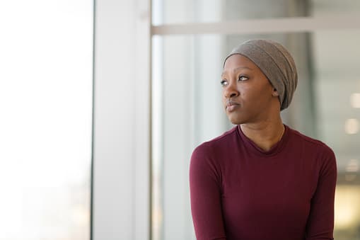 A beautiful woman with cancer wears a hat. She is seated near a large bay of windows. She is taking a break from receiving treatment. The woman is looking out the window with a contemplative expression.
