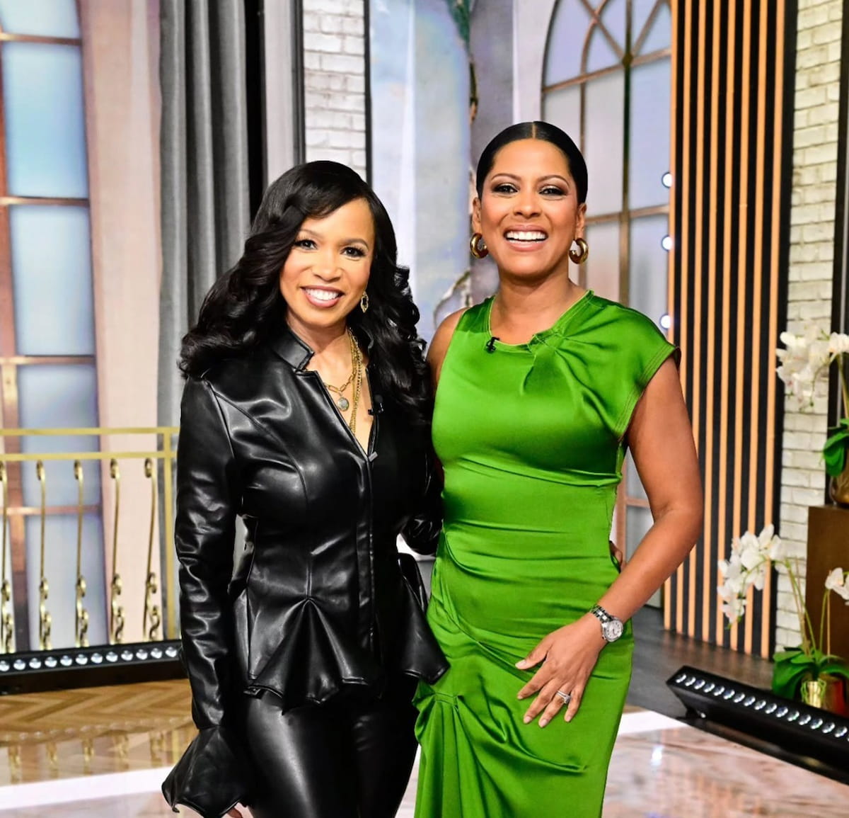 Two smiling Black women posing in a TV studio. The woman on the left wears a black faux leather peplum jacket and pants with gold necklaces. The woman on the right wears a vibrant green satin-like dress.