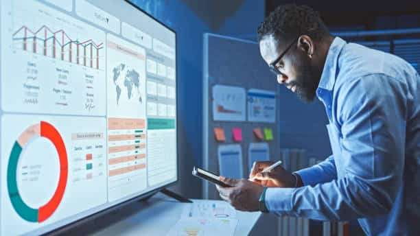 A man with glasses and a beard looks intently at a large monitor displaying a data dashboard with charts, graphs, and a world map, while holding a smartphone and stylus.