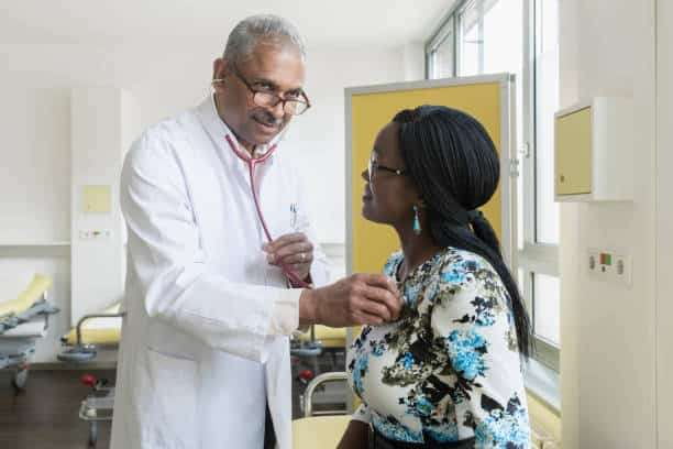 An older male doctor in a white lab coat smiles while using a red stethoscope to listen to the chest of a female patient with dark hair and a floral top.