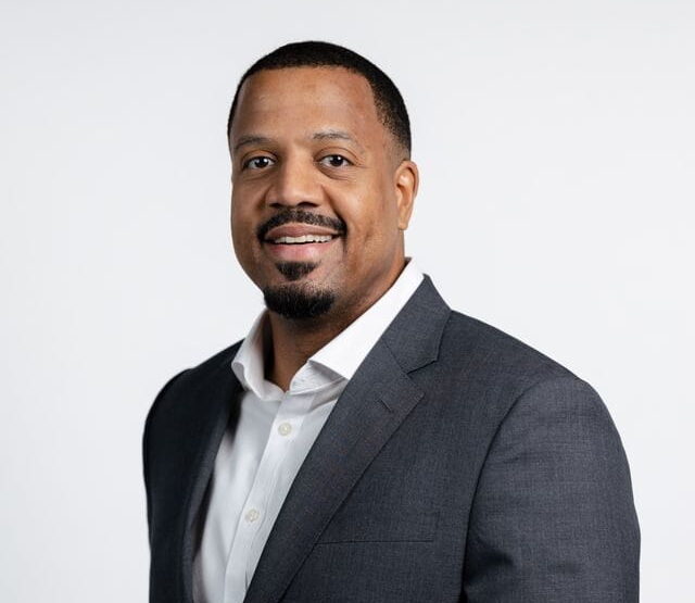 Headshot of a smiling Black man in a dark gray suit jacket and white shirt against a white background.