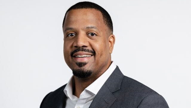 Headshot of a smiling Black man in a dark gray suit jacket and white shirt against a white background.
