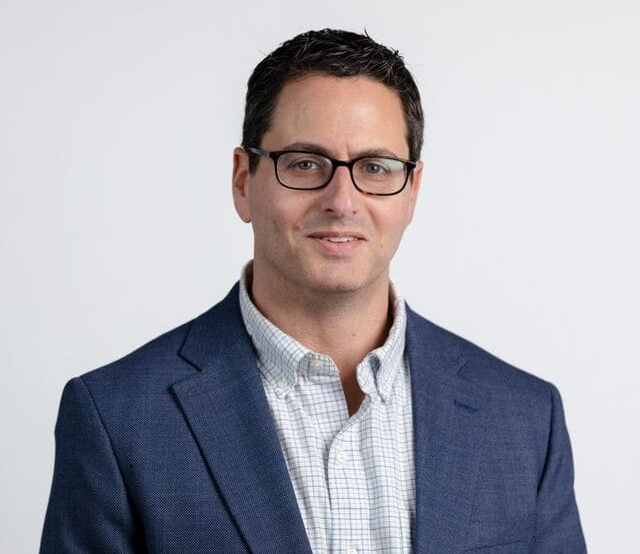 A man with dark hair and glasses wearing a blue blazer and checkered shirt smiles at the camera against a white background.