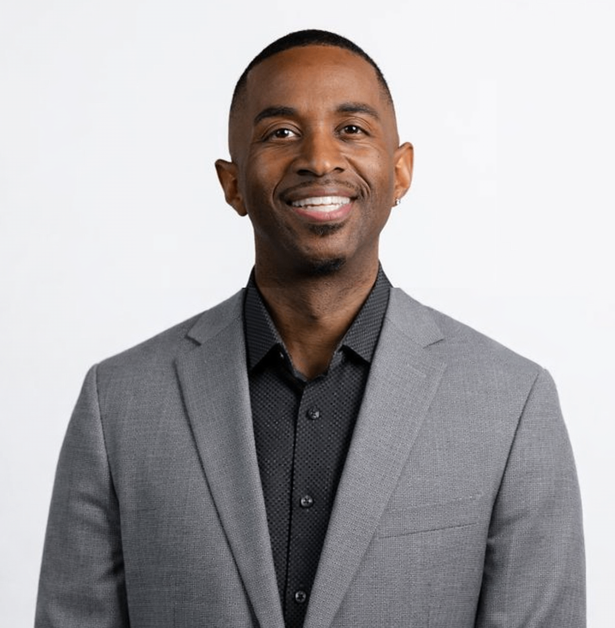 Headshot of a smiling Black man in a gray suit and black shirt against a white background.