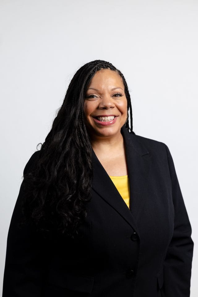 Headshot of a smiling Black woman with long, dark braids, wearing a black blazer over a yellow top, against a white background.