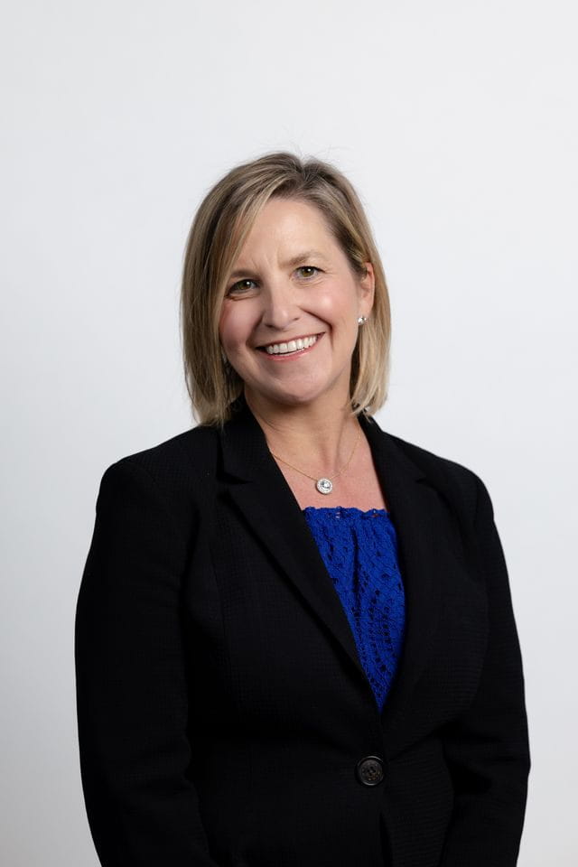 Headshot of a smiling woman with blonde hair, wearing a black blazer and a blue top, against a white background.