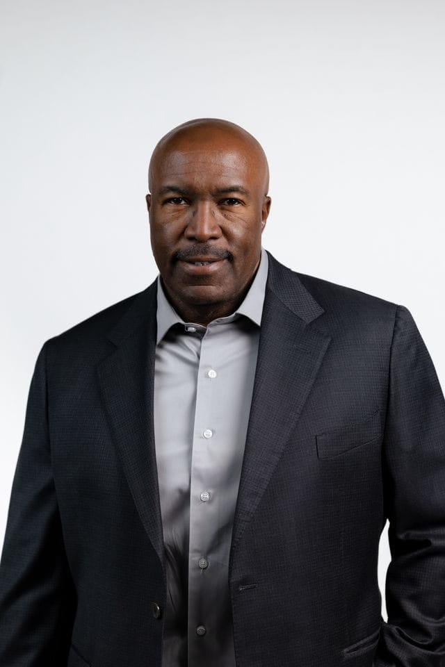 Headshot of a bald, dark-skinned man with a mustache wearing a dark suit jacket and a light gray shirt.