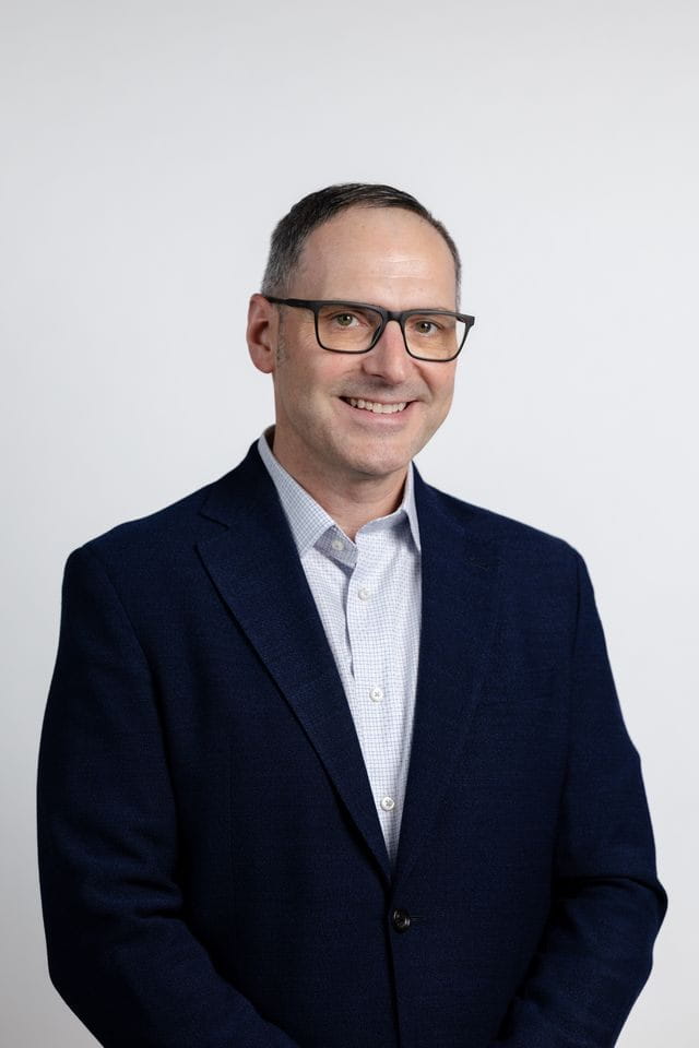 Headshot of a smiling man with short dark hair and black-framed glasses, wearing a blue checkered shirt and a navy blazer.