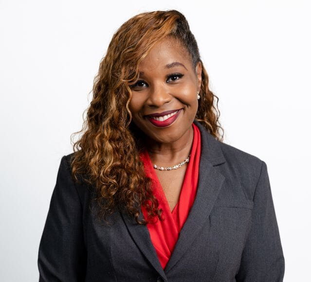 Headshot of a smiling woman with long, wavy brown hair, wearing a dark blazer and red top against a white background.