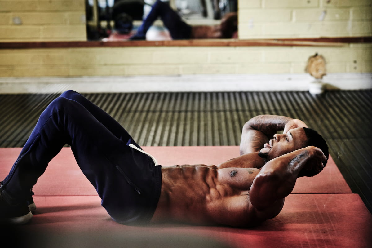 A muscular man with a defined six-pack performs crunches on a red mat in a gym, hands behind his head, eyes closed in focus. A blurred mirror reflection is in the background.