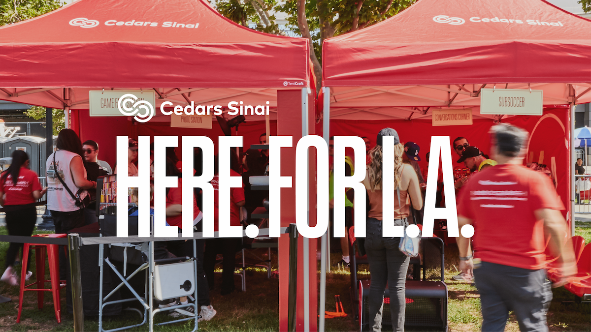 Two red Cedars Sinai tents at an outdoor community event with people interacting. Large white text overlay reads 'HERE. FOR L.A.'