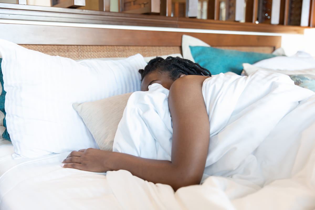 A person with dreadlocks sleeping on their side in a bed, covered by a white duvet, with their face hidden by a pillow.