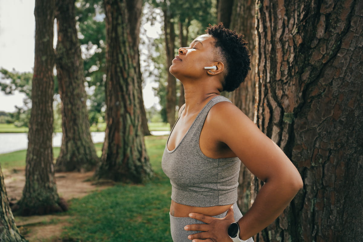 A Black woman with short, curly hair, wearing a grey sports bra and white earbuds, stands with her eyes closed and head tilted back, looking peaceful in a park surrounded by trees.