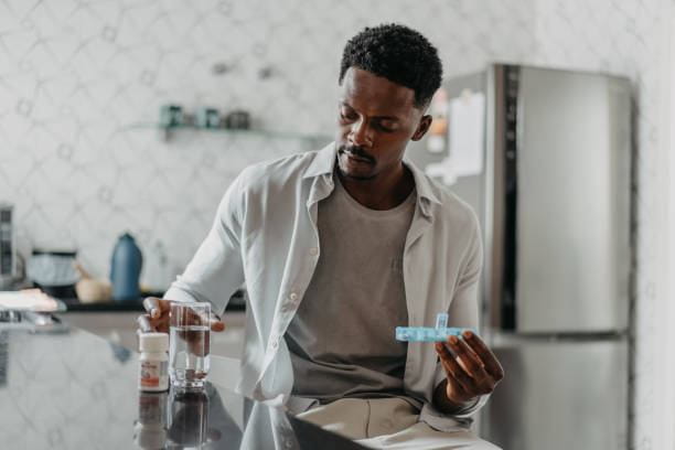A Black man in a kitchen, looking down at a blue pill organizer in his hand, with a glass of water and a pill bottle on the counter beside him.