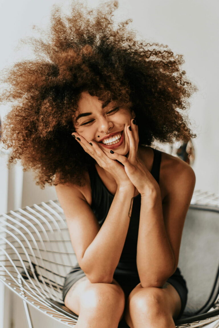 A young woman with voluminous curly hair laughing heartily, eyes closed, with her hands cupping her cheeks. She has a wide smile and nose piercings.