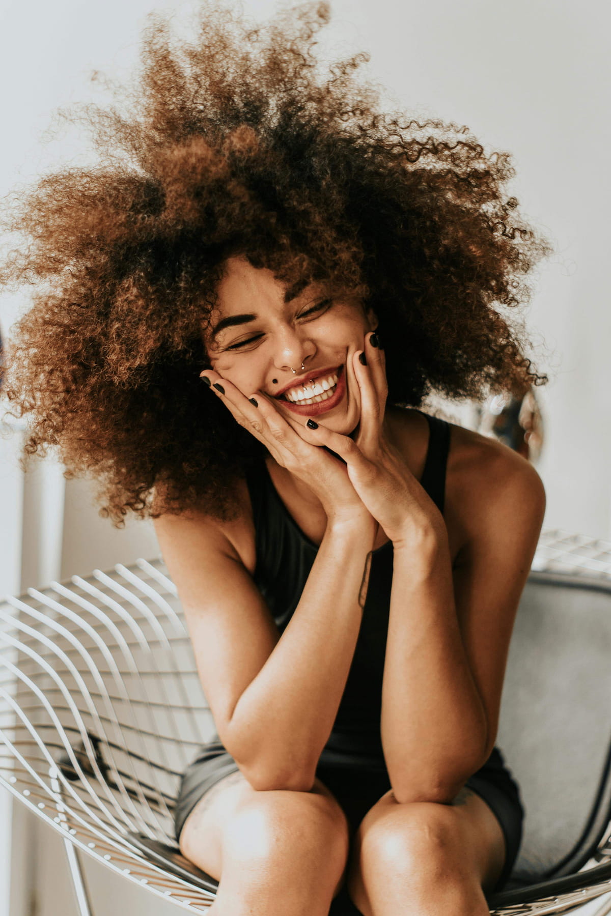 A young woman with voluminous curly hair laughing heartily, eyes closed, with her hands cupping her cheeks. She has a wide smile and nose piercings.