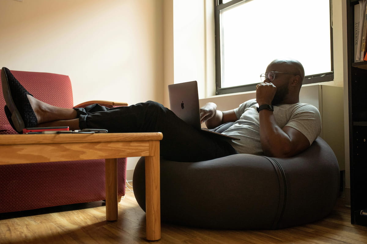 A man with glasses and a beard reclines on a dark bean bag chair, using a laptop on his lap. His feet are propped on a wooden table where two smartphones rest. A bright window is behind him, and a bookshelf is visible on the right.