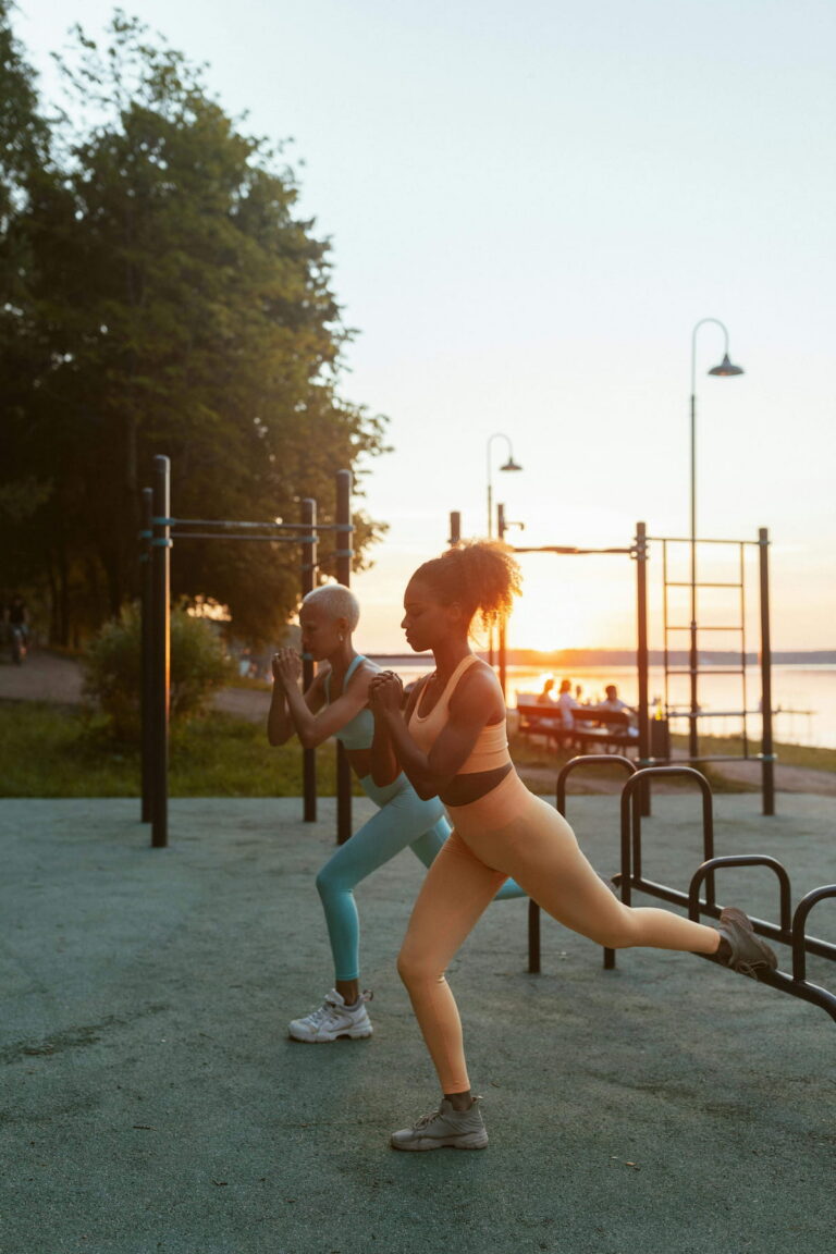 Two women in athletic wear exercising at an outdoor gym during sunrise or sunset. One woman in peach activewear performs a Bulgarian split squat, while another in light blue activewear does a squat. Trees, water, and calisthenics equipment are in the background.