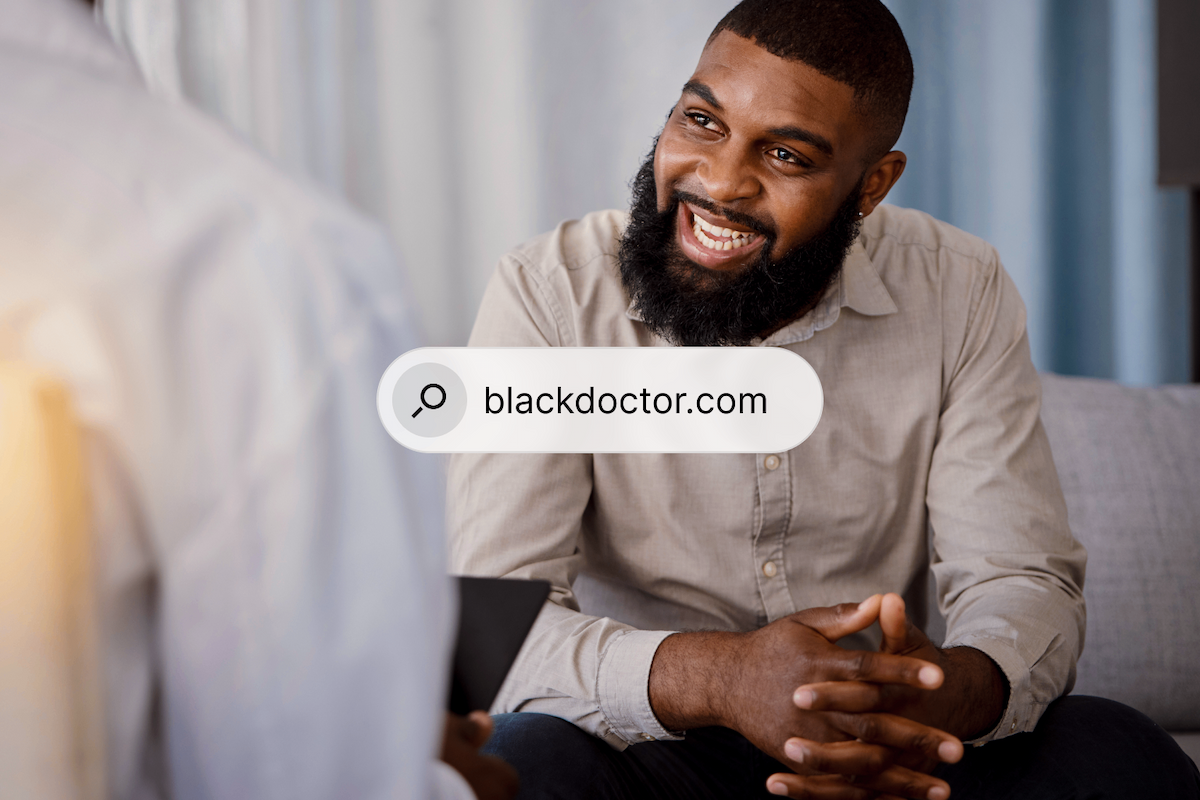 A Black man with a beard smiles broadly while talking to a doctor, whose back is to the viewer. A search bar overlay reads 'blackdoctor.com'.
