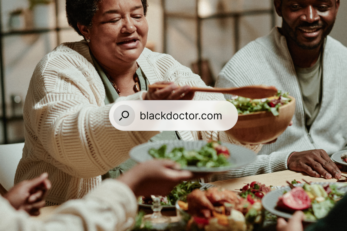 A Black woman serves salad at a family dinner, with a 'blackdoctor.com' search bar overlay.