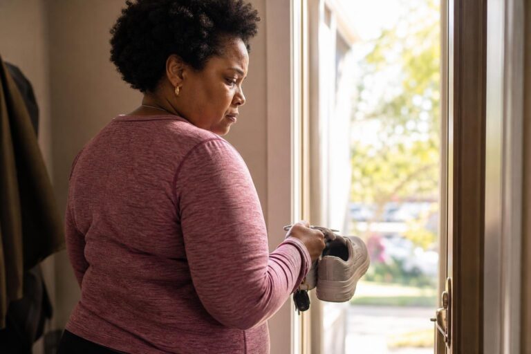 Black woman with ADHD pauses at a doorway holding walking shoes during a weight-control routine.
