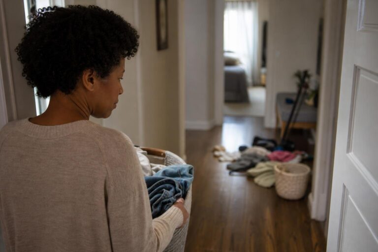 Black woman with ADHD pauses in a doorway beside unfinished laundry, looking concerned.