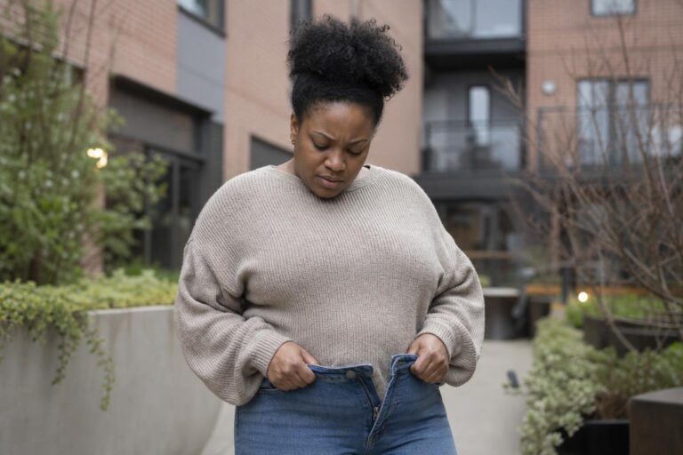 Black woman outdoors looking down while adjusting her jeans, concerned about antidepressant weight gain.