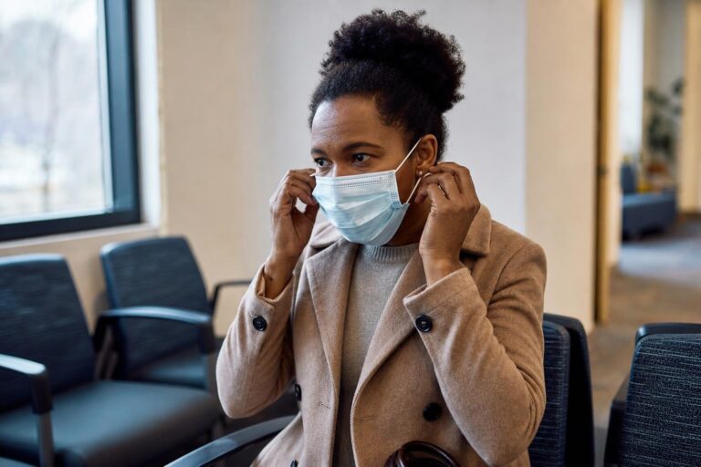 Black mother adjusts her face mask in a clinic waiting area for flu prevention.