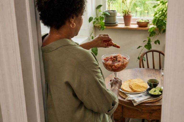 Over-the-shoulder view of a woman serving Mexican-style shrimp cocktail with tortillas in morning light.