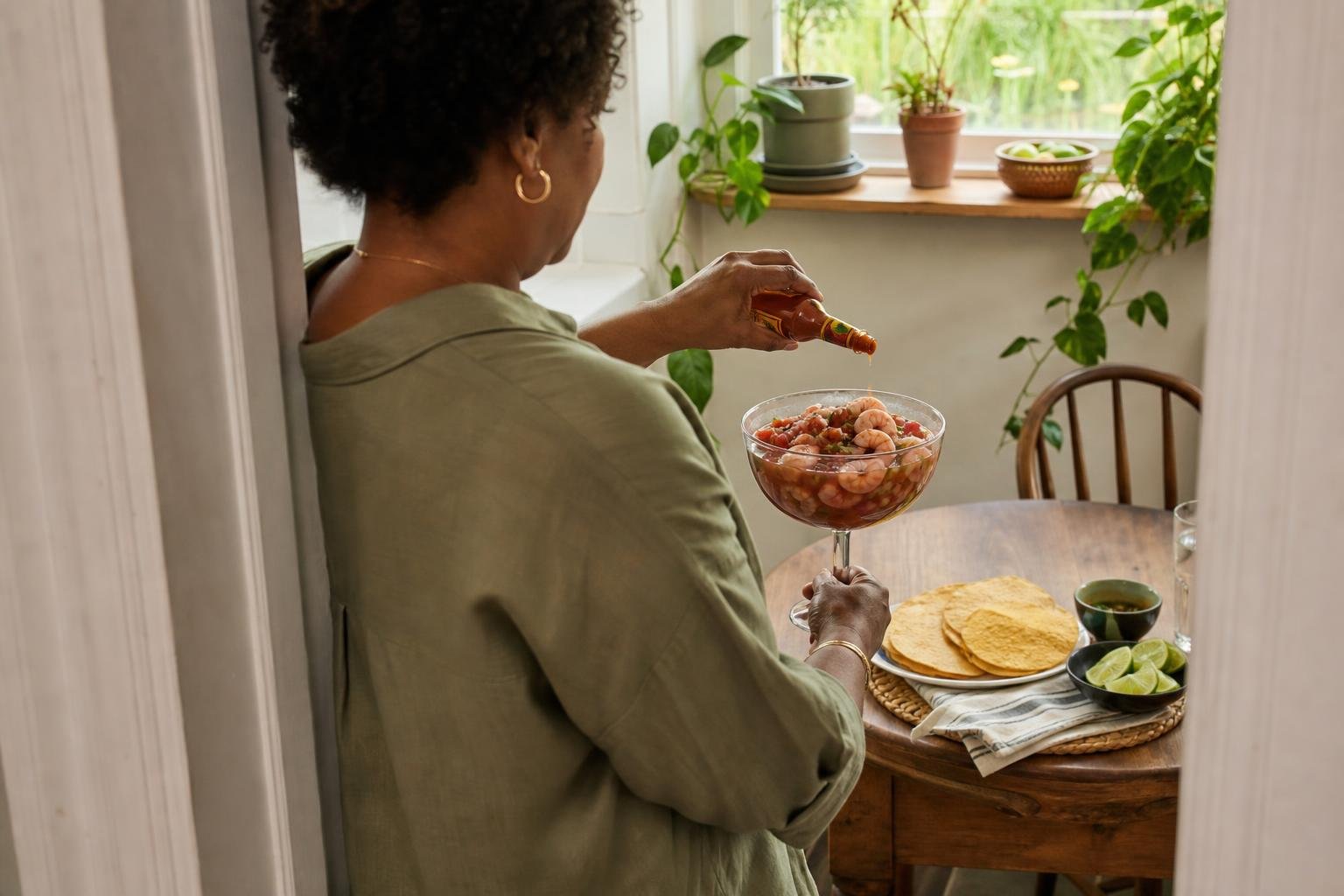 Over-the-shoulder view of a woman serving Mexican-style shrimp cocktail with tortillas in morning light.