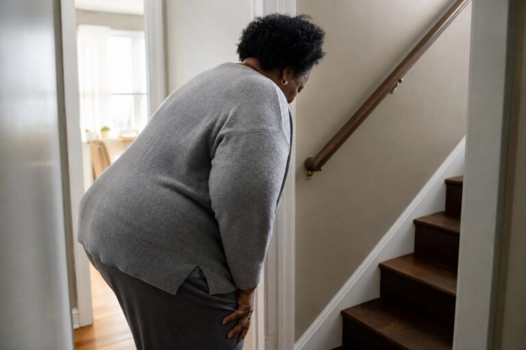 Black woman pauses at stairs with knee pain, reflecting obesity and arthritis concerns.