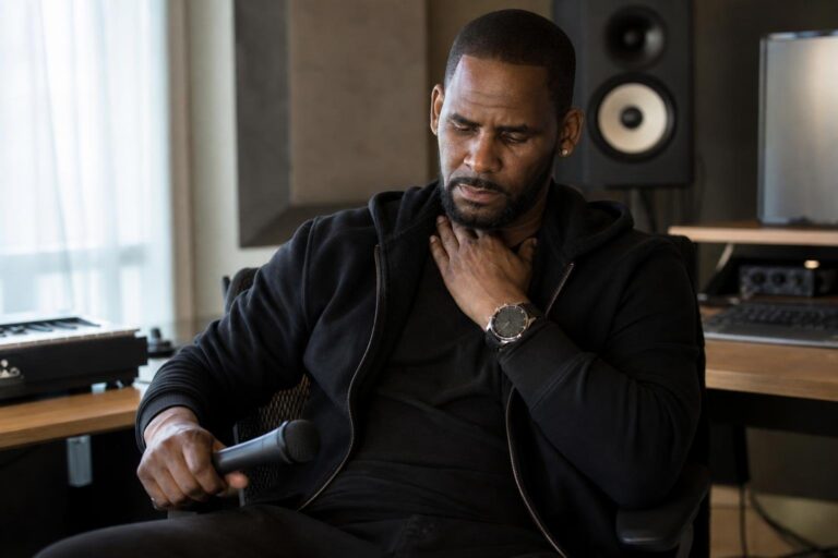 Black male vocalist sets down a microphone at a desk after R. Kelly throat surgery news.
