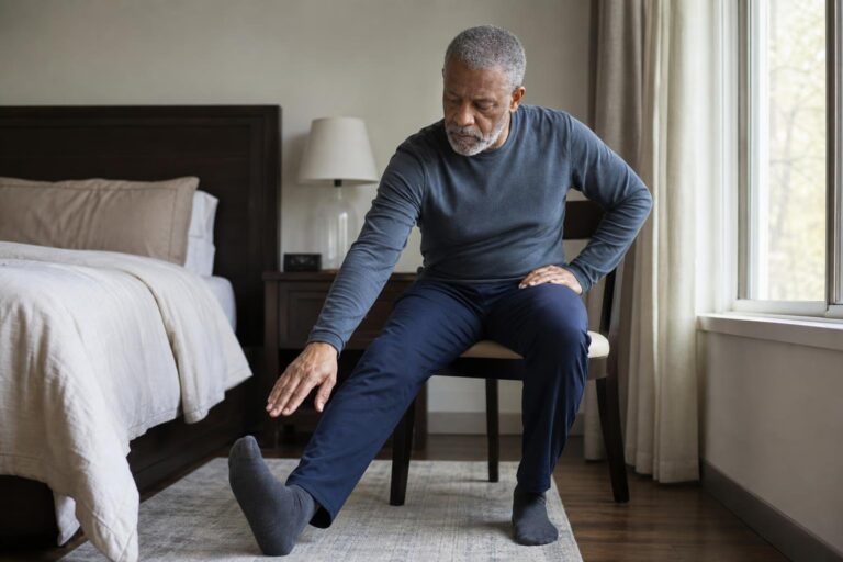 Older man practices a careful seated hamstring stretch beside a bedroom window.